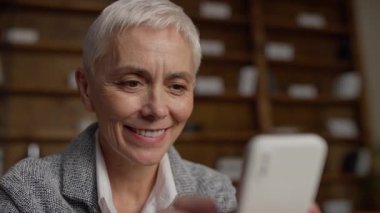 Kısa gri saçlı, gülümseyen, akıllı telefonuyla internetten alışveriş yapan, rahat ve şık bir ev ofisinde oturan olgun bir kadın..