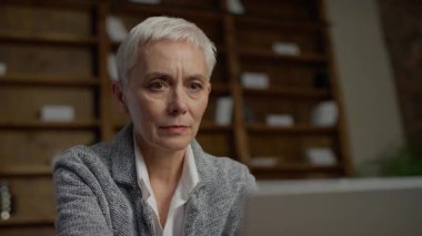 Kısa beyaz saçlı, odaklanmış yaşlı bir kadın, rahat bir evde dizüstü bilgisayarına dikkatlice bakarken çenesini eline dayar..