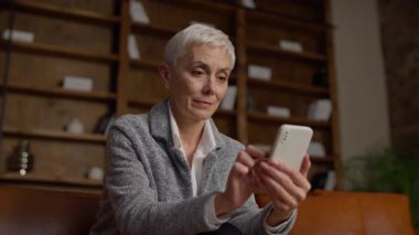 Kısa beyaz saçlı yaşlı bir kadın arka planda kitaplıklarla rahat bir oturma odasında otururken akıllı telefonuna bakar..