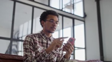 Gözlüklü ve ekose gömlekli genç adam akıllı telefonuyla kayarken şok olmuş görünüyor. Modern bir ev ortamında beklenmedik kötü haberlere tepki veriyor..