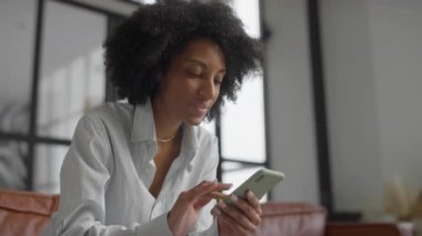 Modern bir oturma odasında akıllı telefonuyla online alışveriş uygulamasına göz atan genç bir kadın..