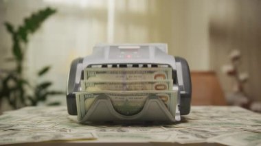 İyi ışıklandırılmış bir odada, nakit parayla çevrili, para sayma makinesinin, yığınla Amerikan doları banknotlarını işlerken dramatik bir şekilde çekilen görüntüsü..
