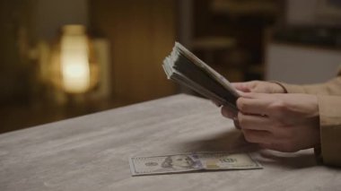 Bir kişi, bir masanın üzerinde, loş ve sıcak bir iç ayarda, ek banknotlar yayılmış, kalın bir yığın Amerikan doları sayar..