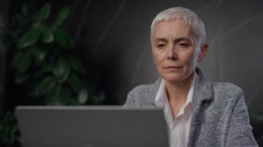 Kısa beyaz saçlı kıdemli bir iş kadını koyu renk dekoru ve yeşil bitkileri olan modern bir ofiste dizüstü bilgisayarına dikkatle bakar..