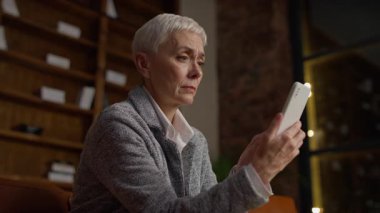 Üst düzey bir kadın akıllı telefonunu incelerken şüpheli görünür, muhtemelen online alışverişe, sosyal medya paylaşımına veya dijital içeriğe tepki verir