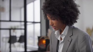 Gri takım elbiseli profesyonel bir kadın, geniş pencereli ve şömineli modern bir ofiste laptopuyla çalışıyor..