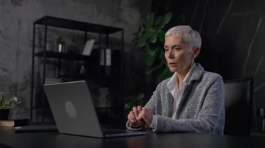 Üst düzey profesyonel bir kadın karanlık bir ofis masasında oturuyor, bilgisayarında sanal bir toplantı yapıyor, endişeli ve tartışmaya odaklanmış görünüyor..