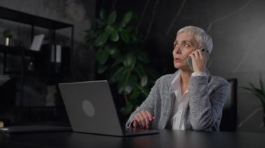 Üst düzey profesyonel bir kadın karanlık bir ofiste oturuyor. Bilgisayarı üzerinde çalışırken ciddi bir telefon görüşmesi yapıyor. Odaklanmış ve endişeli görünüyor..