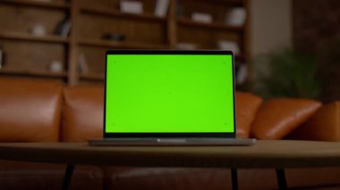 Kahverengi deri kanepesi ve kitaplığı olan, rahat bir oturma odasında, ahşap bir masaya yerleştirilmiş, yeşil ekranlı modern bir laptop..