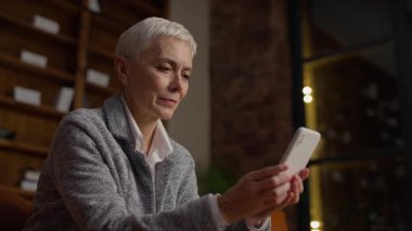 Elinde akıllı bir telefonla gülümseyen olgun bir kadın kitap rafı arka planında rahat bir ofiste otururken olumlu bir mesajın tadını çıkarıyor..