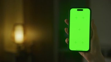 Özelleştirme için yeşil ekranlı akıllı telefonu tutan kişi, loş ışıklandırılmış arka plan ve sıcak bir lamba ile donatılmış..