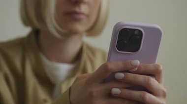 Akıllı telefonuyla mesaj atan bir kadın, elinde şık, mor bir çanta olan bir cihaz tutarken, yumuşak ışıklı bir ortamda evde oturuyor..
