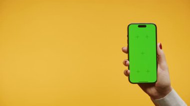 Kırmızı ojeli bir kadın, yeşil ekranlı akıllı bir telefonu canlı sarı bir arka plana karşı tutuyor..