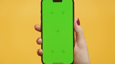 Kırmızı tırnak cilası olan bir kadın elinde yeşil ekranlı akıllı bir telefon tutuyor, canlı sarı bir arka plana zumlanmış bir şekilde gösteriliyor..