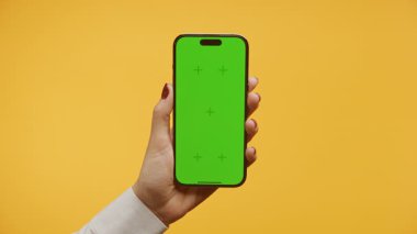 Kırmızı ojeli bir kadın elinde yeşil ekranlı akıllı bir telefon, koyu sarı bir arka planda..