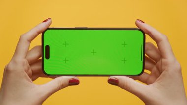 Kırmızı ojeli bir kadın elinde akıllı bir telefon, yeşil ekranlı, parlak sarı bir arka planda..