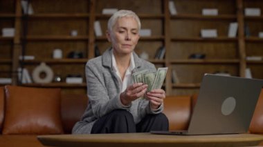 Olgun bir kadın ev ofisinde oturmuş, dikkatle Amerikan dolarlarını sayıyor, açık bir dizüstü bilgisayarın yanında, odaklanmış bir ifadeyle..