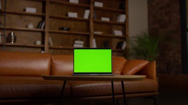 Kitap rafları ve deri mobilyalarla dolu, ev ofisinin ahşap masasında yeşil ekranlı bir dizüstü bilgisayarın sinemadaki görüntüsü..