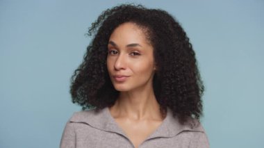 Kıvırcık siyah saçlı neşeli bir kadının portresi, neşeyle göz kırpıyor ve sırıtıyor, kendine güvenini ve çekiciliğini mavi bir arka plana yansıtıyor..