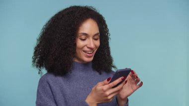 Kıvırcık siyah saçlı neşeli bir kadının portresi, gözleri açık ve ağzı açık, mavi arka planda akıllı telefonuna bakarken heyecanla ve şaşkınlıkla tepki veriyor..