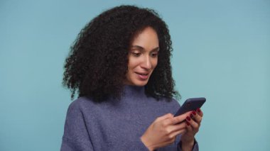 Kıvırcık siyah saçlı neşeli bir kadının portresi. Akıllı telefonuna bakarken heyecandan yumruğunu sıkıyor. Başarısını veya başarısını mavi arka planda kutluyor..