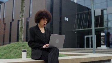 Siyah takım elbiseli neşeli bir iş kadını modern bir ofis binasının dışındaki dizüstü bilgisayarına bakarken heyecandan yumruğunu sıkıyor..