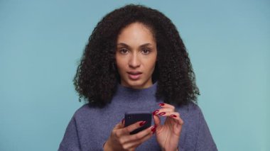 Mavi kazaklı genç bir kadın online alışveriş yaparken akıllı telefonuna şüpheli bir ifadeyle bakıyor, satın alma seçeneğini değerlendiriyor..