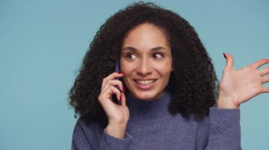 Mavi kazaklı neşeli bir kadın heyecanlı bir şekilde telefonuyla konuşuyor, gülümsüyor ve mavi bir arka planda el kol hareketi yapıyor..