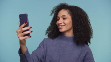 Mavi kazaklı neşeli bir kadın telefonuyla video konuşmasında gülümsüyor, mavi arka planda sanal bir sohbete girişiyor..