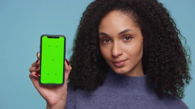Mavi kazaklı, kendine güvenen bir kadın yeşil ekranlı bir akıllı telefonu elinde tutuyor ve mavi arka planda kişiselleştirme için boş bir görüntü sunuyor..