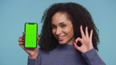 Mavi kazaklı neşeli bir kadın yeşil ekranlı bir akıllı telefonu kaldırırken, iyi bir jest yapıyor, özelleştirme için boş bir görüntü sergiliyor, mavi arka planda yer alıyor..