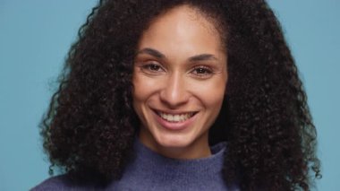 Kıvırcık saçlı neşeli bir kadın yakın plan bir portrede sıcacık gülümsüyor, mavi bir süveter giyiyor ve mavi bir arka planı var..