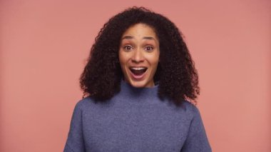Kıvırcık saçlı neşeli bir kadın şaşkınlık ve heyecan ifadesi sergiliyor. Ağzı açık pembe bir arka plana bakıyor..