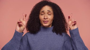 Kıvırcık saçlı bir kadın her iki elinde de parmaklarını çapraz yapar, gözlerini kapatır ve pembe arka planda beliren umut, beklenti veya hüsnükuruntu ifade eder.