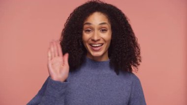 Kıvırcık saçlı neşeli bir kadın, parlak bir gülümseme ile kameraya el sallıyor. Selamlama, dostluk ya da pembe arka planda heyecan ifade ediyor..