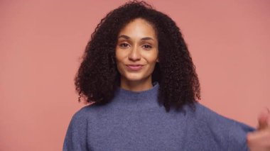 Kıvırcık saçlı neşeli bir kadın onay verirken sıcak bir şekilde gülümser. Onayını, pozitifliğini ve pembe bir arka plana karşı cesaretini gösterir..