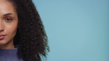 Kıvırcık saçlı, mavi kazaklı, yumuşak mavi arka planlı bir kadının yakın plan portresi, yüzünün yarısı görünür..