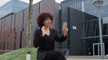 Siyah takım elbiseli neşeli bir iş kadını modern bir ofisin önünde oturuyor, akıllı telefonuyla video görüşmesi yaparken el sallıyor ve gülümsüyor..