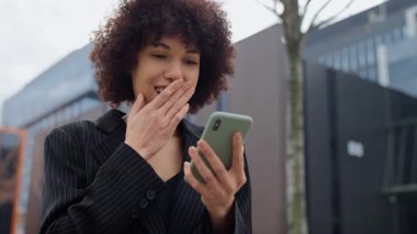 Siyah takım elbiseli neşeli bir iş kadını, bir akıllı telefonu tutarken yumruğunu kaldırarak büyük bir başarı için heyecanını dile getiriyor..