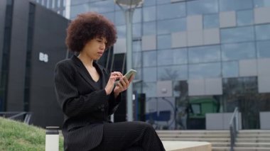 Siyah takım elbiseli profesyonel bir kadın modern bir ofisin önünde duruyor, akıllı telefonuyla online bir alışveriş uygulamasına bakarken konsantre oluyor..