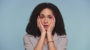 Kıvırcık saçlı genç bir kadın şaşkınlık içinde yüzünü tutuyor. Beklenmedik haberlere şaşırmış bir ifadeyle karşılık veriyor..