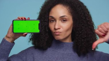 Kıvırcık saçlı genç bir kadın, yeşil ekranlı akıllı bir telefonu tutarken baş parmağını kaldırma hareketi göstererek onaylamayan bir ifade takınır..