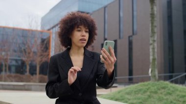 Siyah çizgili takım elbiseli profesyonel bir kadın şehir ortamında oturuyor, akıllı telefonuyla bir video görüşmesi yapıyor, odak noktasıyla iletişim kuruyor..