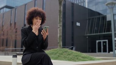 Siyah çizgili takım elbiseli neşeli bir iş kadını, akıllı telefonuna bakarken heyecanla tepki veriyor. Modern bir şehirde, açık havada kutlamada yumruğunu kaldırıyor..