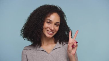 Kıvırcık saçlı, gri bluzlu neşeli bir kadın, mavi bir arka plana karşı pozitifliğini ve dostluğunu ifade ederek parmaklarıyla barış işareti yapıyor..