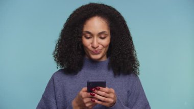 Mavi kazak giyen kıvırcık saçlı neşeli bir kadın akıllı telefonuna bakar. Mavi arka plandaki bir mesaja verdiği yanıtı düşünceli bir şekilde düşünür..