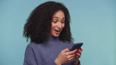 Mavi kazak giyen kıvırcık saçlı neşeli bir kadın akıllı telefonuna bakarken heyecanla tepki veriyor. Mavi arka planda iyi haberi kutluyor..