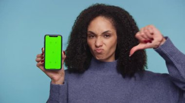 Kıvırcık saçlı, mavi kazak giyen hoşnutsuz bir kadın yeşil ekranlı bir akıllı telefonu tutarken mavi arka plandaki memnuniyetsizliğini dile getiriyor..