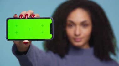 Mavi kazak giyen kıvırcık saçlı bir kadın kameraya doğru yeşil ekranlı bir akıllı telefon tutuyor ve mavi arka plan karşısında kişiselleştirilebilir bir görüntü sağlıyor..