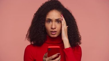 Kırmızı kazaklı bir kadın, pembe bir arka plana güvenmeyerek, şok ve endişe içinde, alnına dokunarak akıllı bir telefon tutuyor..